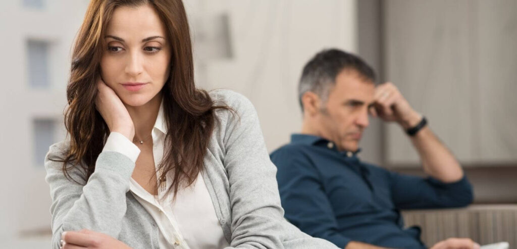 A couple sits on a couch, both looking upset. She is facing towards the camera and he is facing away