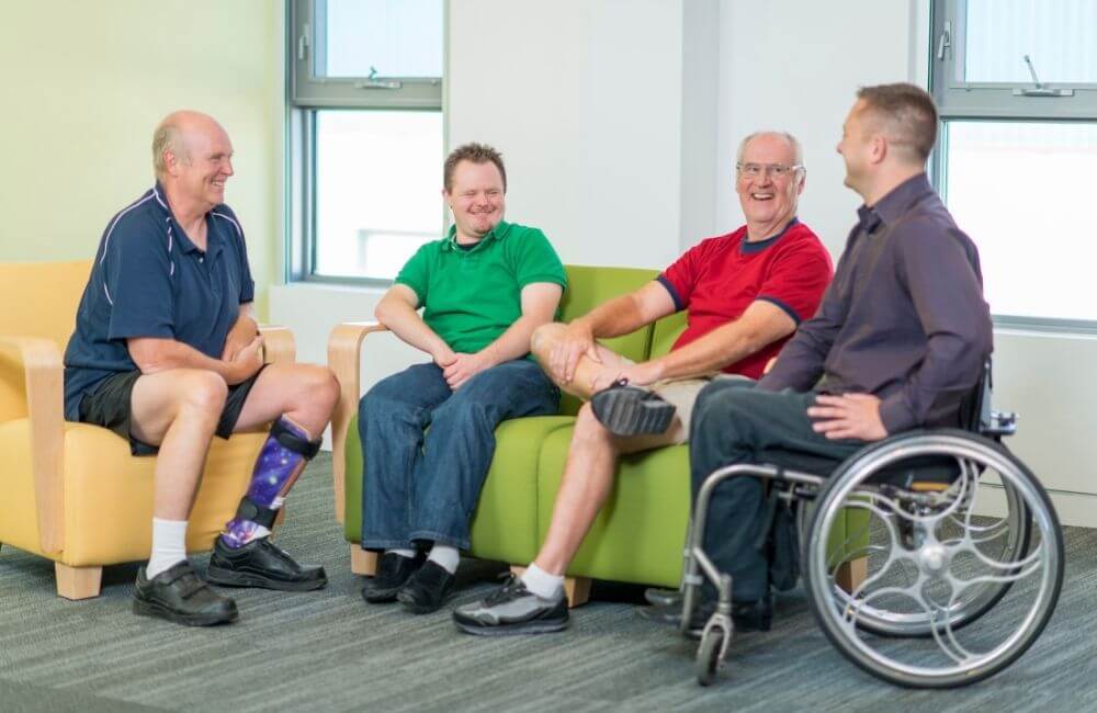 Four men are visiting, one in a purple shirt is in a wheelchair. One is sitting on a yellow chair and two share a green couch