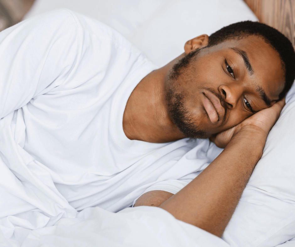 A person with dark skin, a beard, and a short haircut lies awake in a bed fittied with white linens.
