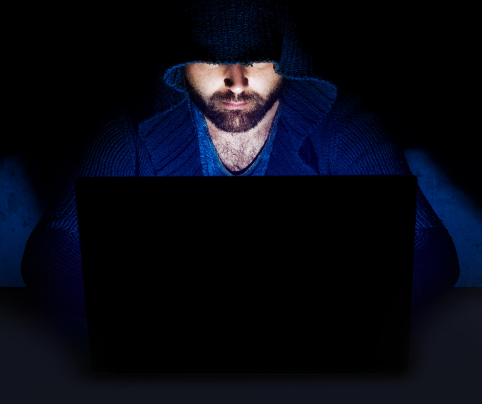 A person with pale skin and a dark brown beard looks at a computer in a dark room with a hood over his head.