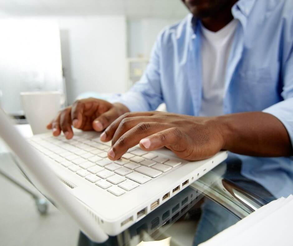 a black man in a blue button up shirt sits down typing on a white laptop computer