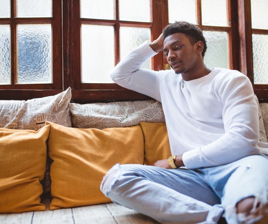 A man with darker skin sits on a couch with cream and yellow pillows. He looks forlorn and lonely. He is wearing a long sleeve white shirt and light denim jeans with a hole in the knee.