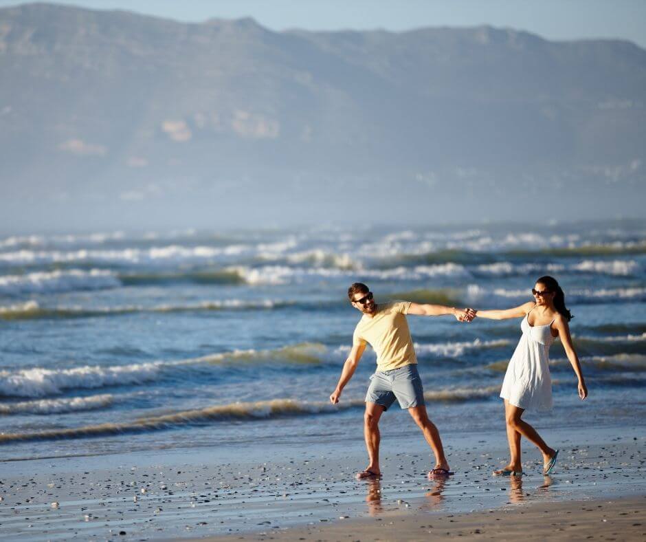 A couple on the beach, walking in the surf, playfully pulling each other by their hands. 