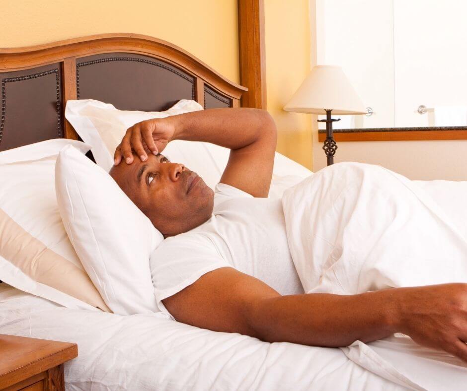 A man with dark skin lies in a bed with all white linens. His hand is to his forehead, signaling that he is having a hard time sleeping