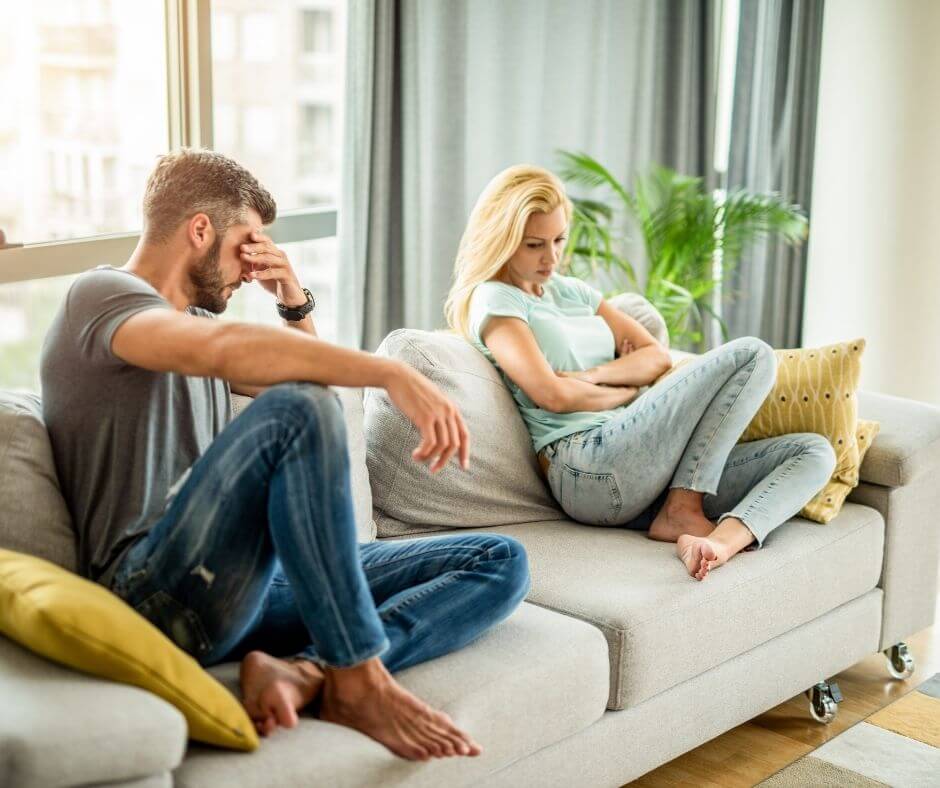 A frustrated man sits on the left side of the couch, his leg bent and his arm resting on his knee, his other hand to his face. A woman with blond hair sits on the other side of the couch with her body pulled away, arms crossed, looking down.