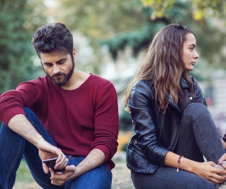 A man with dark hair and beard, wearing a red sweater sits with his back to a woman. She has dark hair and is wearing dark clothing. They are avoiding each other.