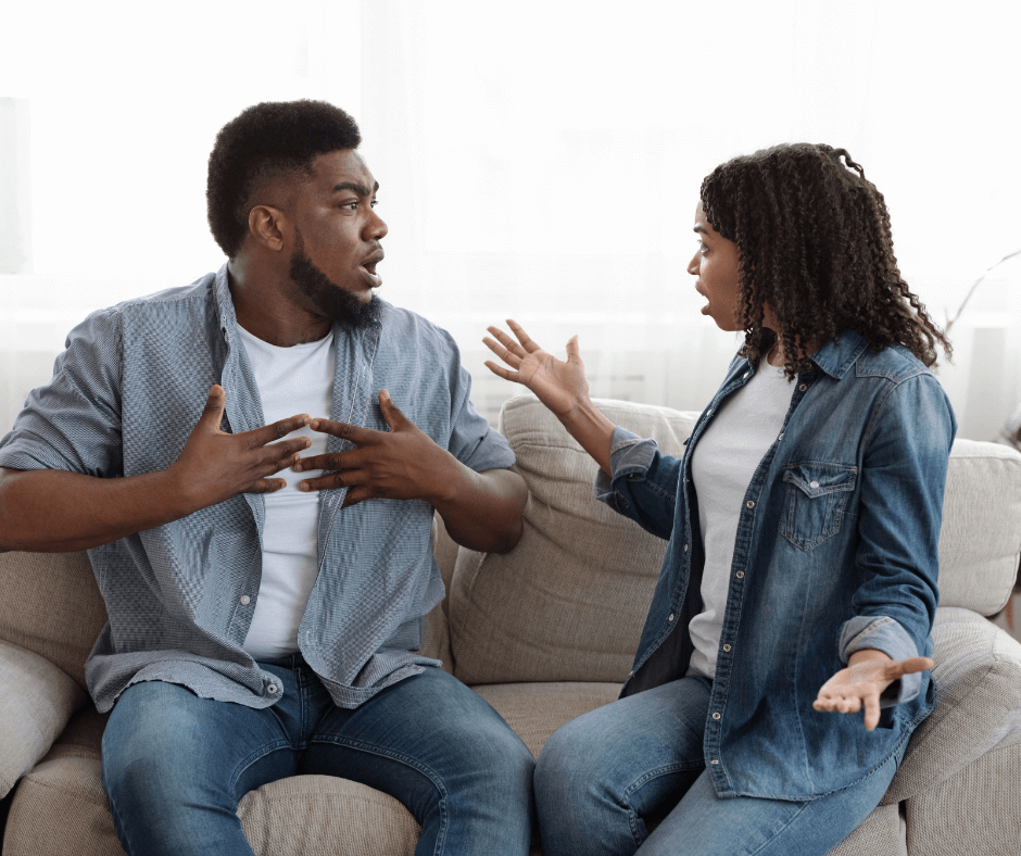 A couple, both with dark skin wearing denim, are sitting on a couch. They are clearly in conflict with one another. 