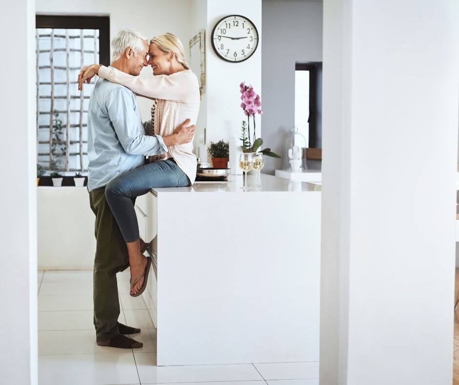 An older woman sits on the kitchen counter while an older man embraces her waist. She has her arms over his shoulders and they're heads are pressed together. 