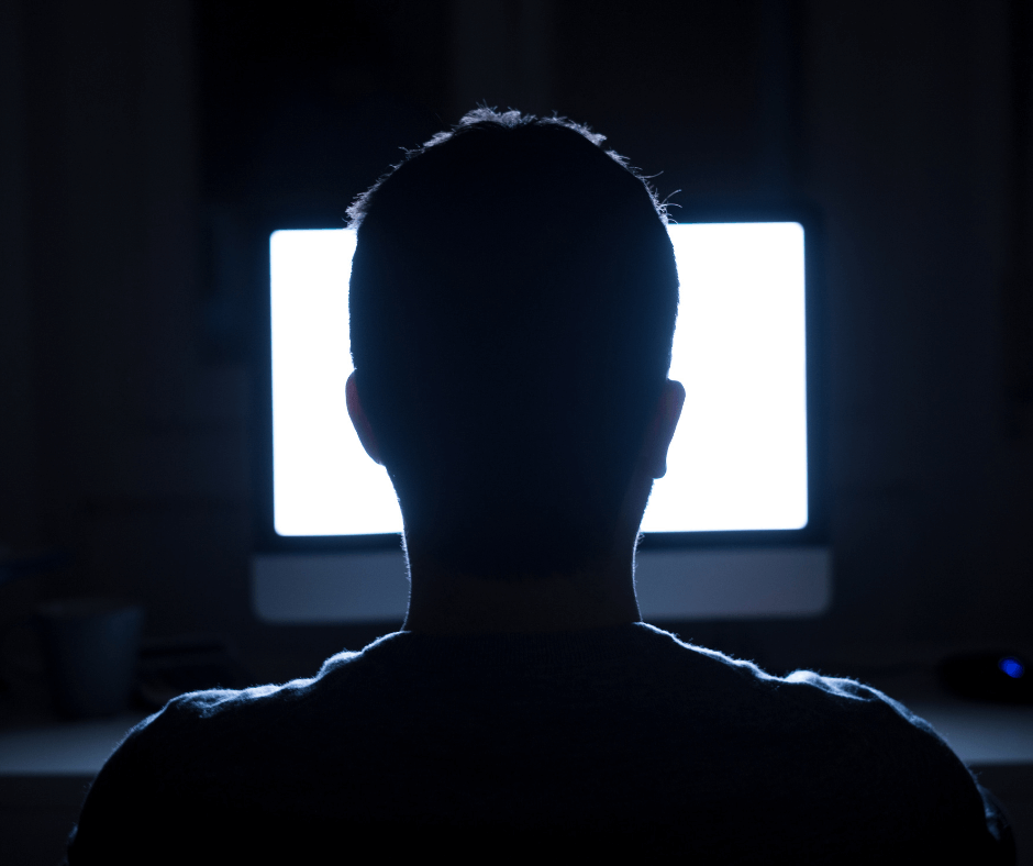 From behind, we see a person staring into a computer screen in a dark room