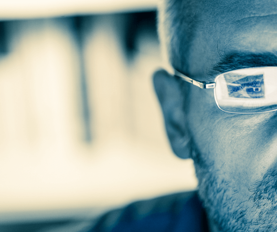 A person with light skin and a beard stares straight ahead with a computer screen reflecting in his glasses