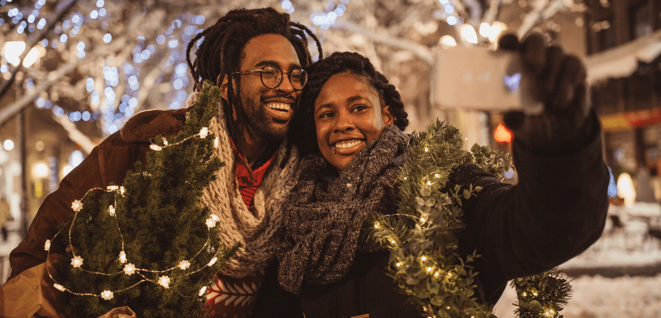An african american couple a male and female stand outdoors holding a christmas tree taking a selfie with their phone, they look very happy and stand close together