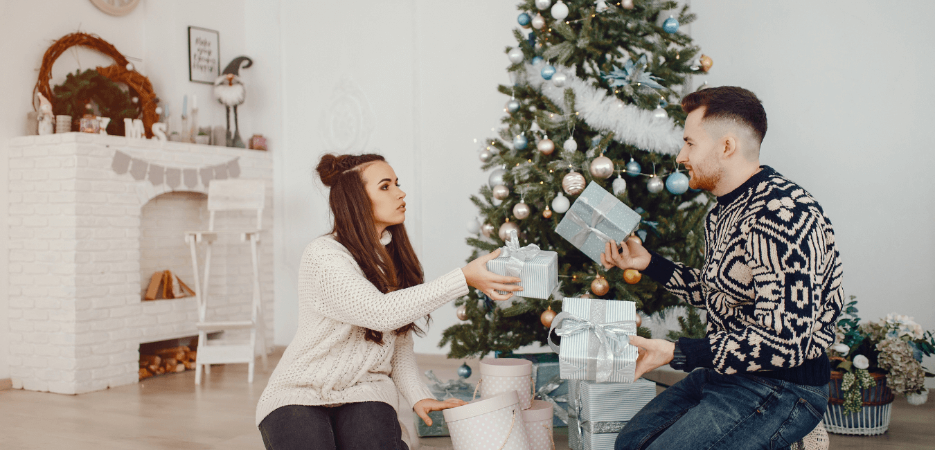 A male and female couple sit on the ground in front of a christmas tree, they are looking at each other while they hold presents, her face looks concerned.