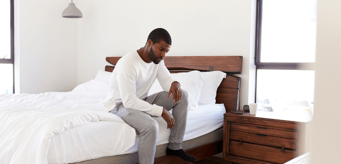 A man looking unhappy sitting on side of his bed in in a broad daylight