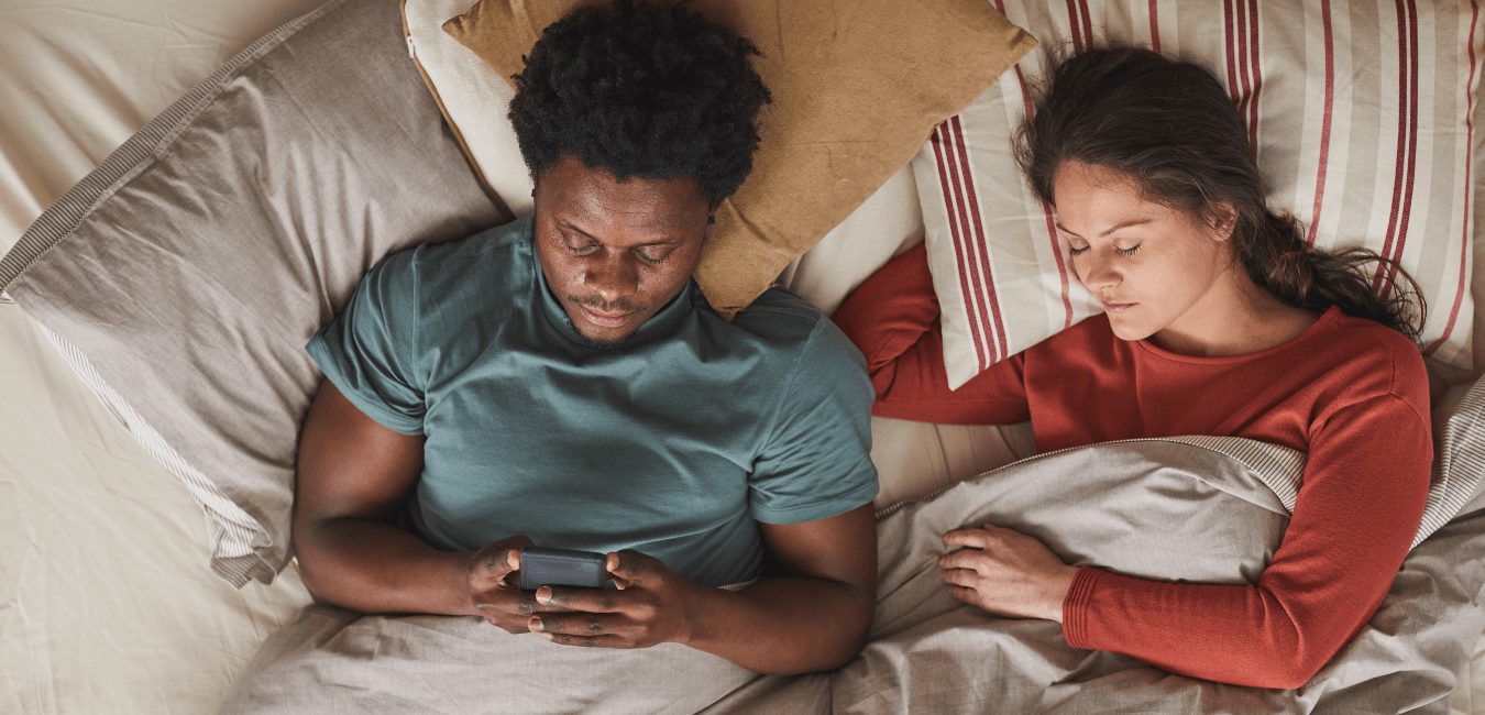 Interracial couple lying on bed and man using his phone while woman is sleeping
