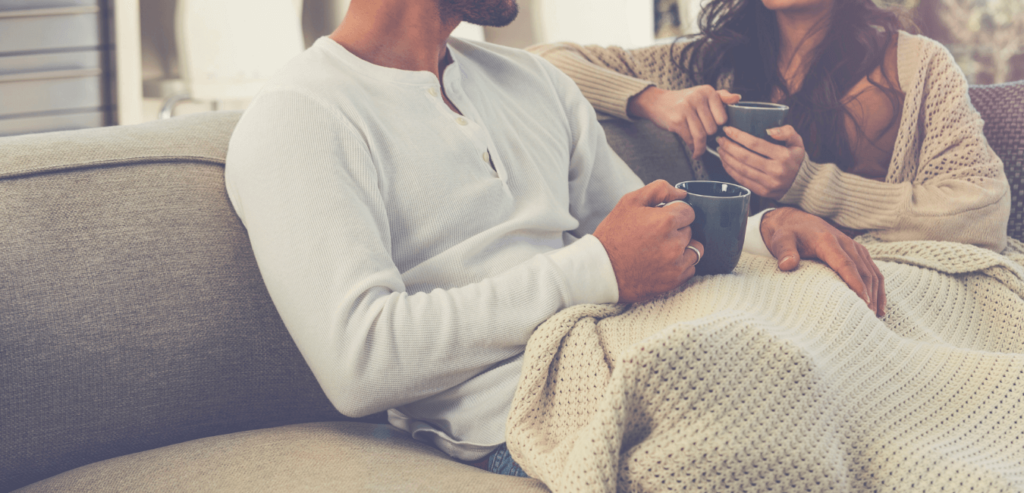 Couple sitting on sofa holding a mug wearing sweater and blanket on their lap getting cozy