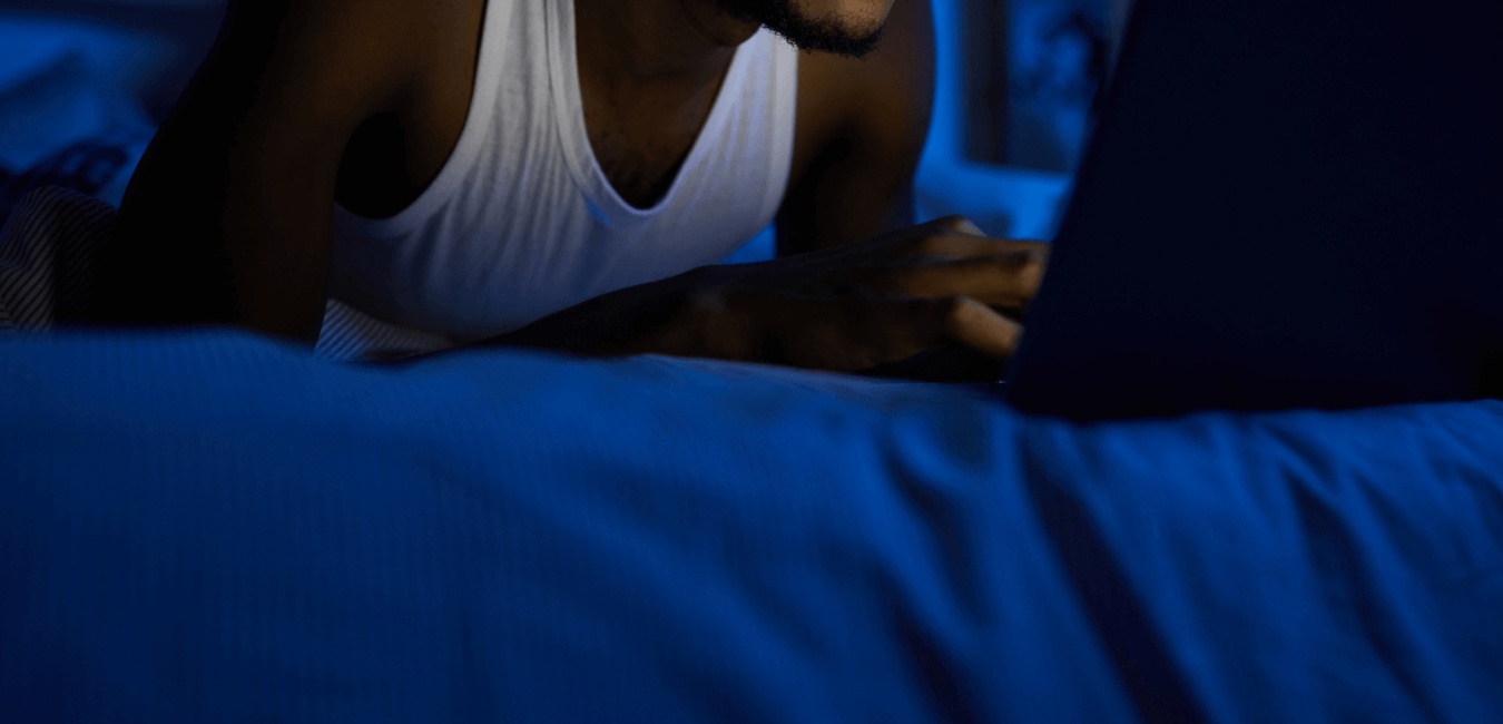 African man wearing white sando on his bed with laptop