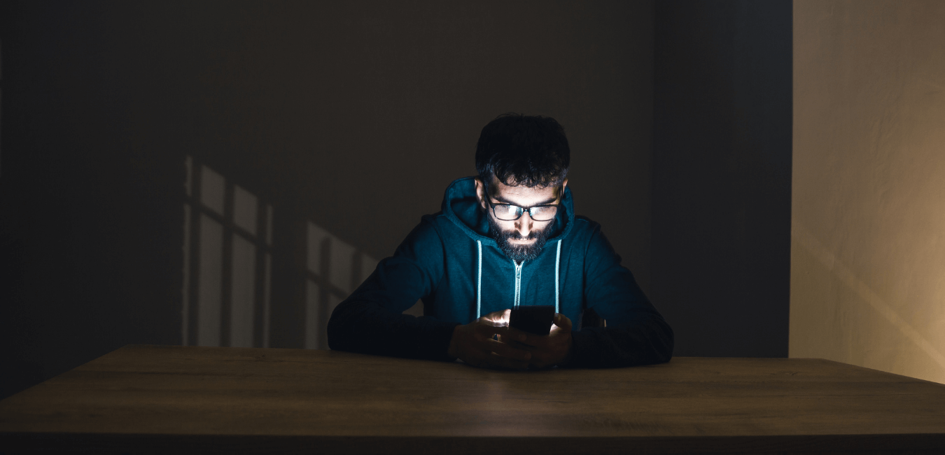 Young man with beard wearing jacket with hood using his phone in a dark room on table
