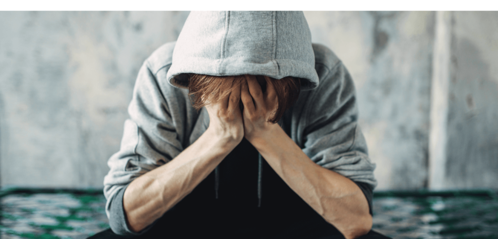 Man wearing brown hoodie sitting near wall looking depressed with his hands on his face