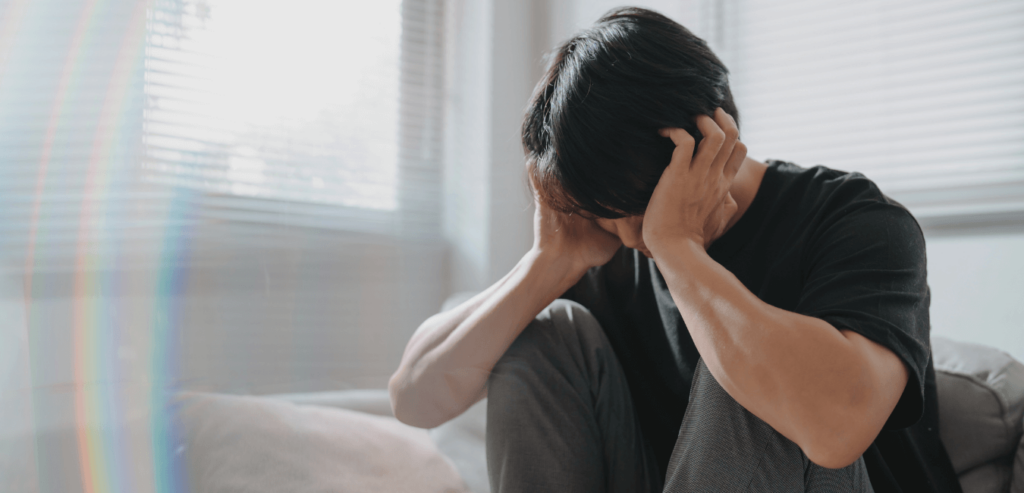 Man sitting on couch near window with his head down and hand on his ear looking depressed