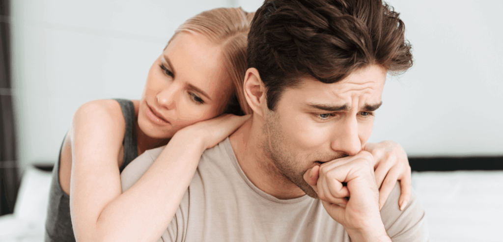 Couple sitting on a bed, wife hugging and comforting her crying husband.