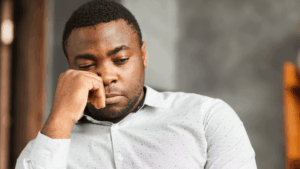 A man of color sits alone in the corner, deep in thought, his hand resting on his face.