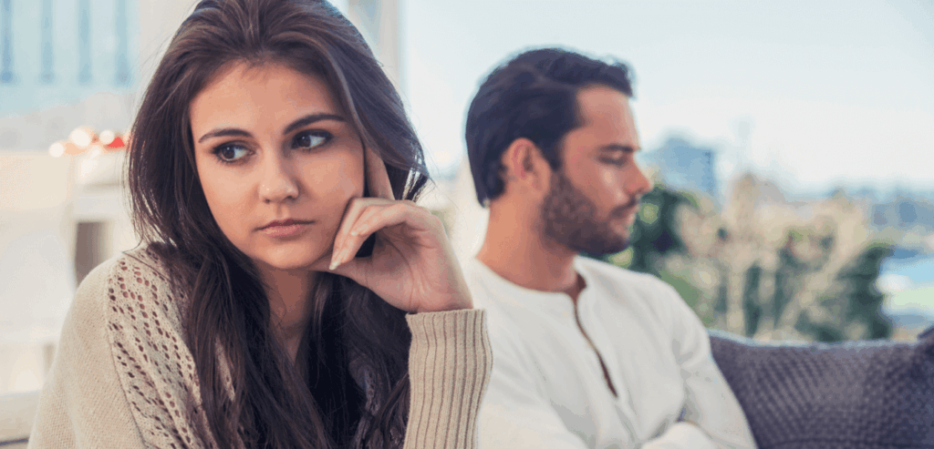 Young couple sits quietly on the couch, not talking or looking at each other, as if something is wrong.
