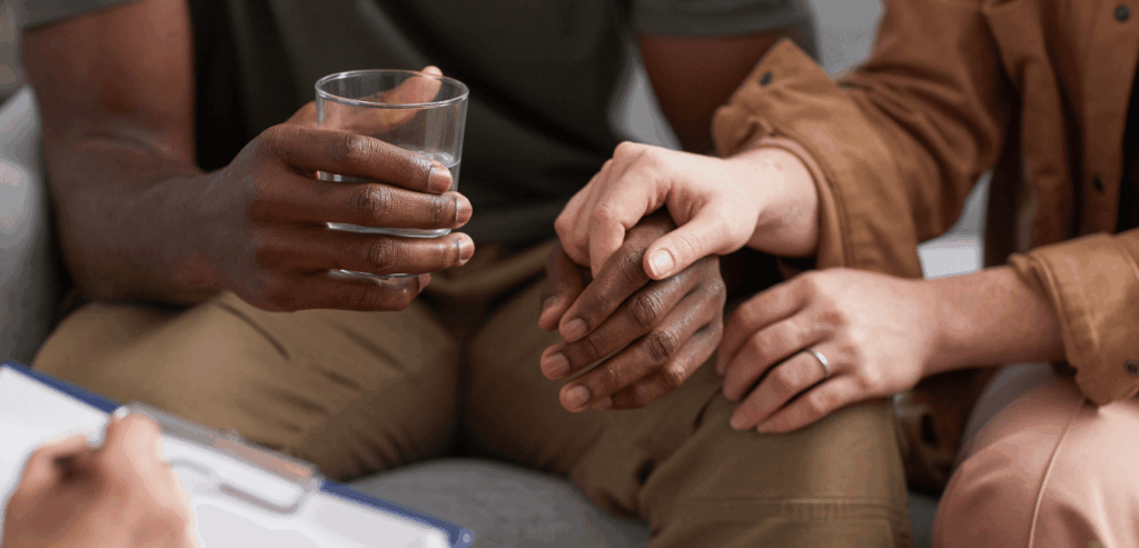 A couple sits on a couch, the man holding a glass of water, while their therapist listens and takes notes.