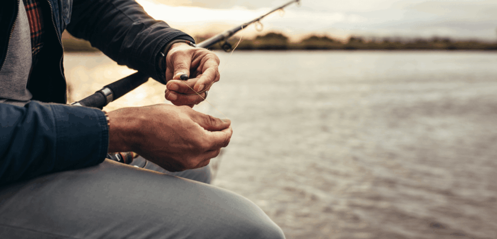 Man sits quietly on the shore, setting up his fishing rod as the sun rises over calm waters