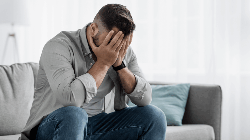 Man sits in silence on his couch, head lowered, hand on his face, battling feelings of shame and addiction.