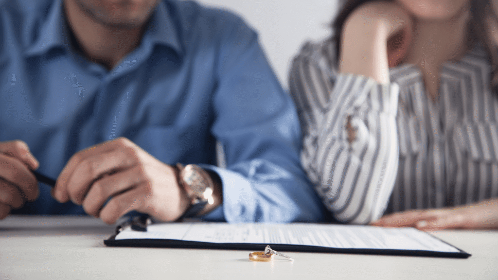 Close-up of wedding ring and divorce papers, couple blurred in background.