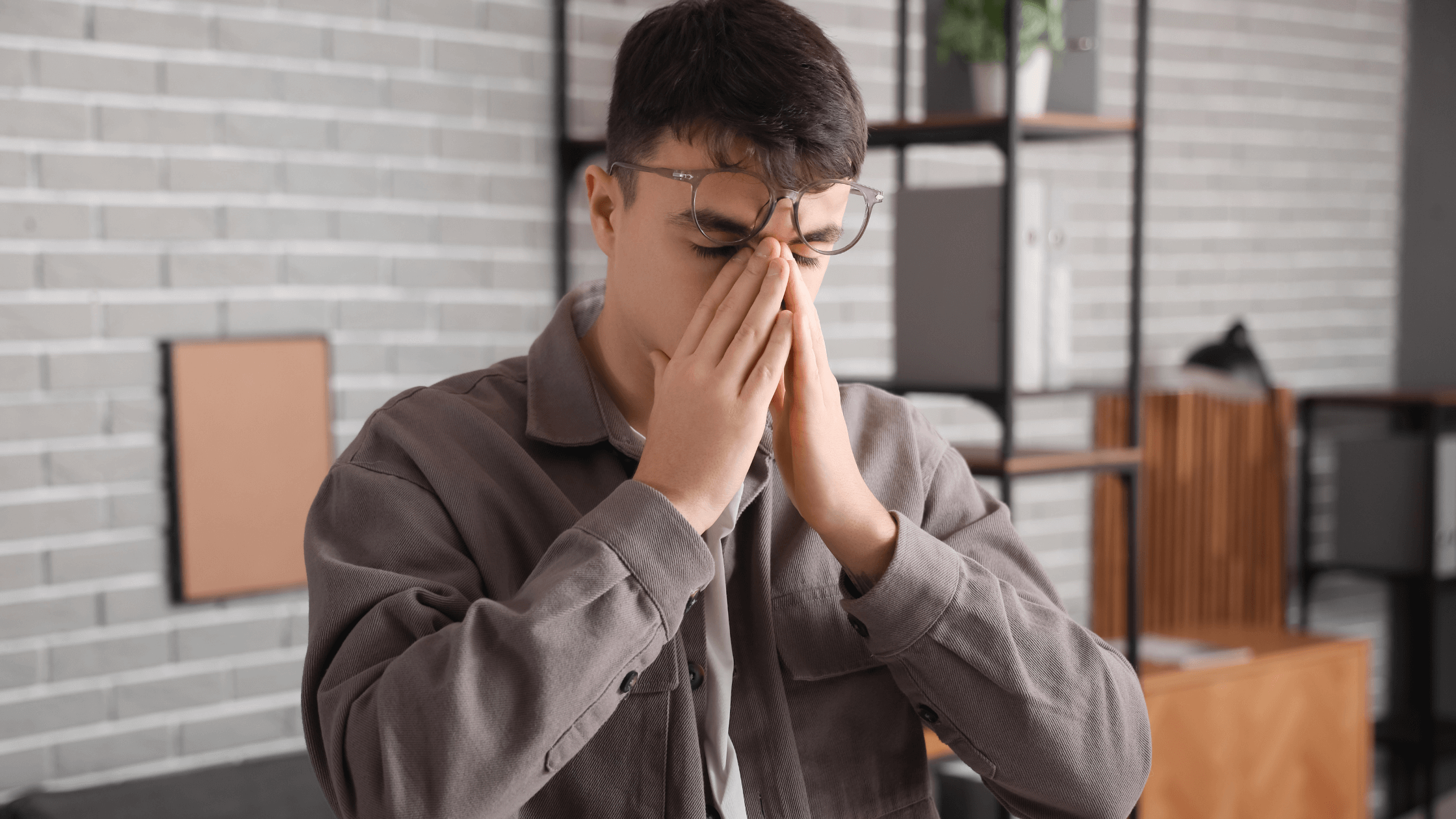 Young man with glasses, eyes closed, hand on face, looking sad and stressed