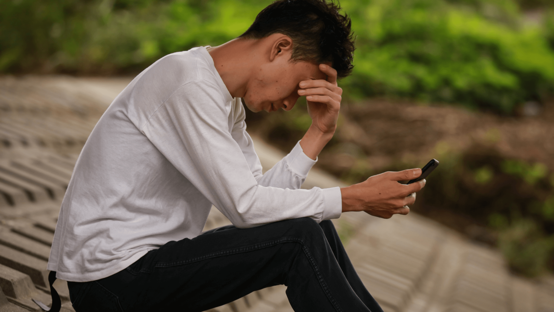 Man sitting outdoors, looking down at phone, hand resting on head.