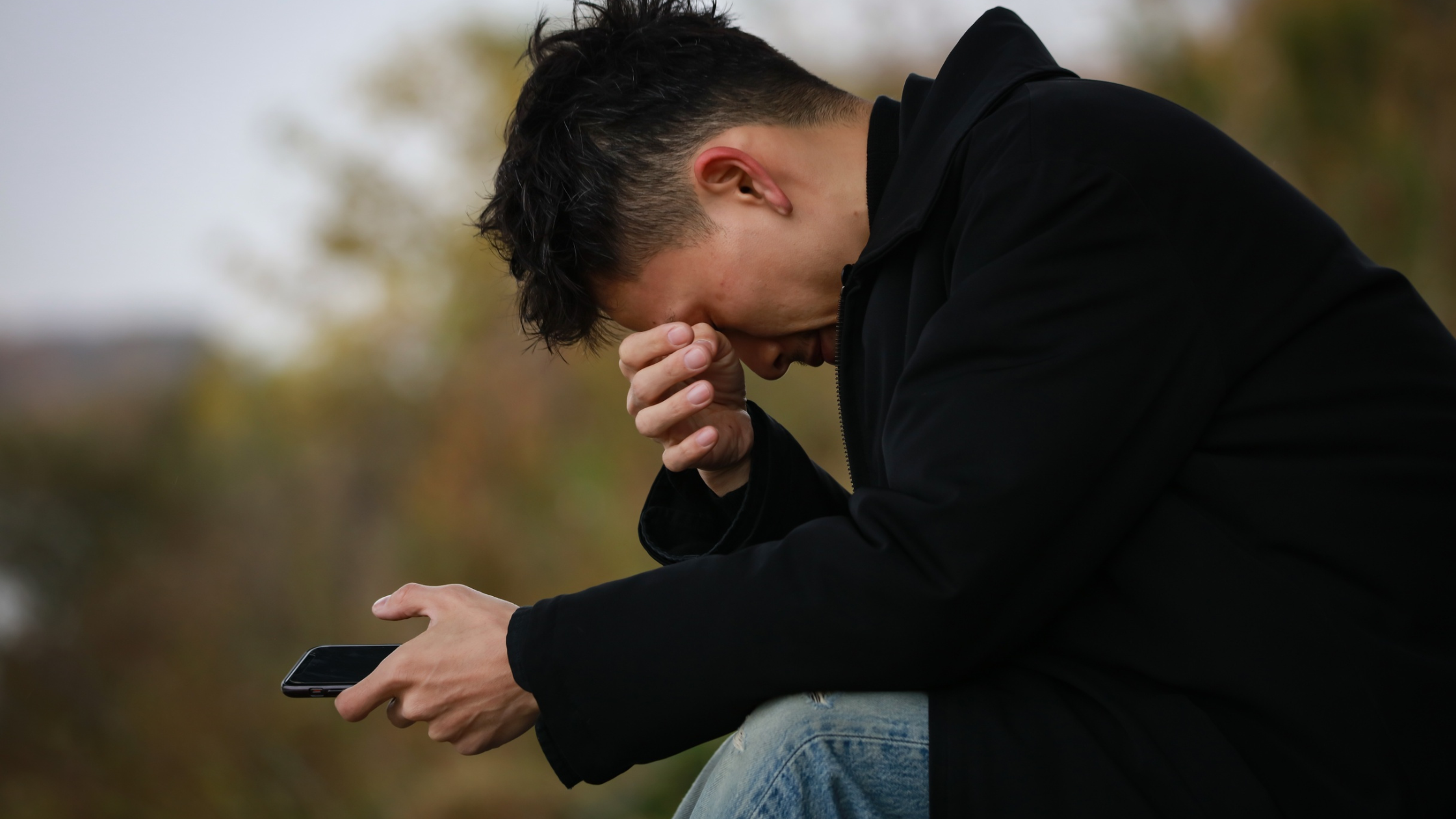 Man sitting outside, holding phone, wiping eyes with hand