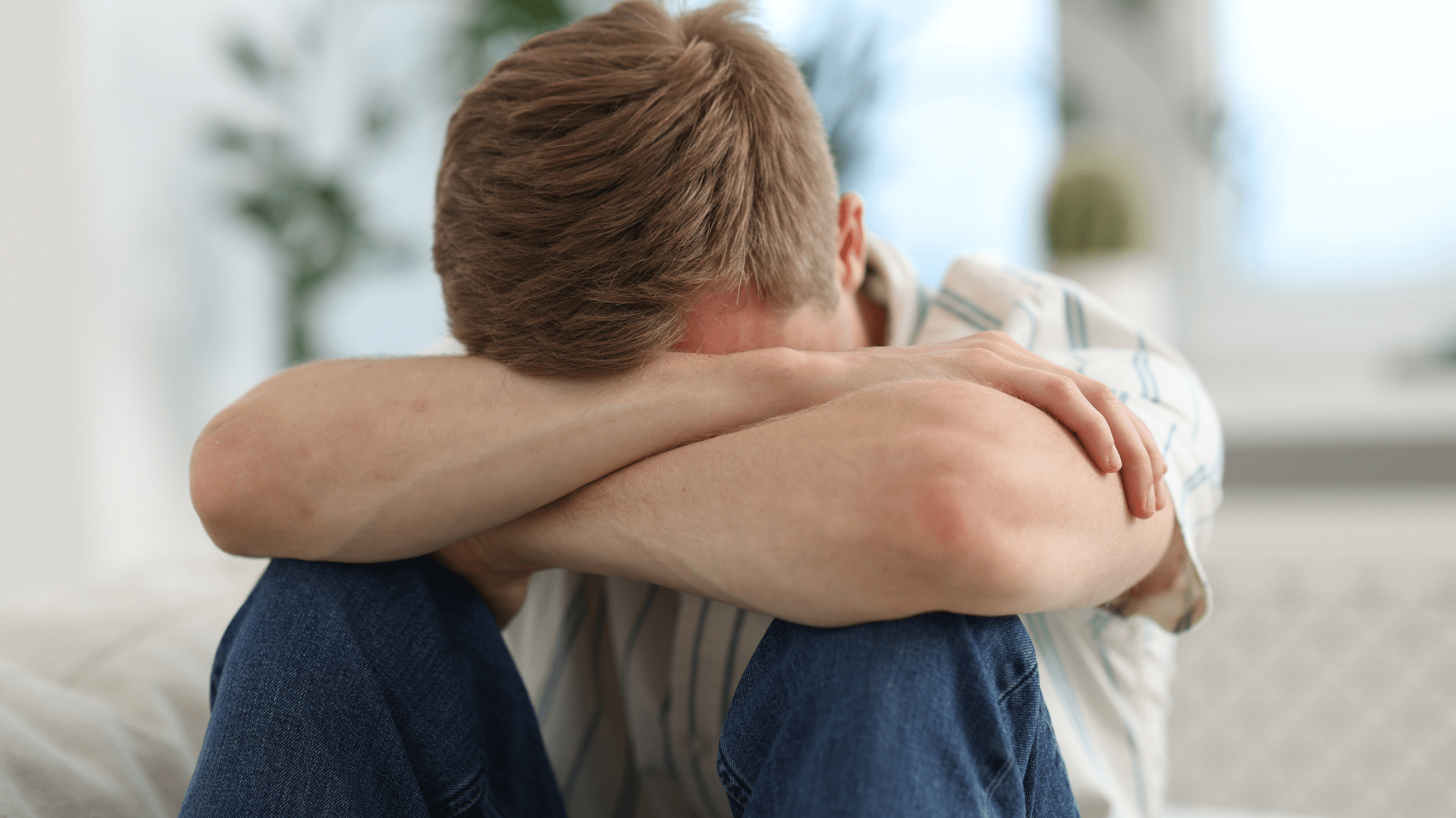 Young man sitting at home with arms crossed over his face, looking like he's experiencing a difficult moment
