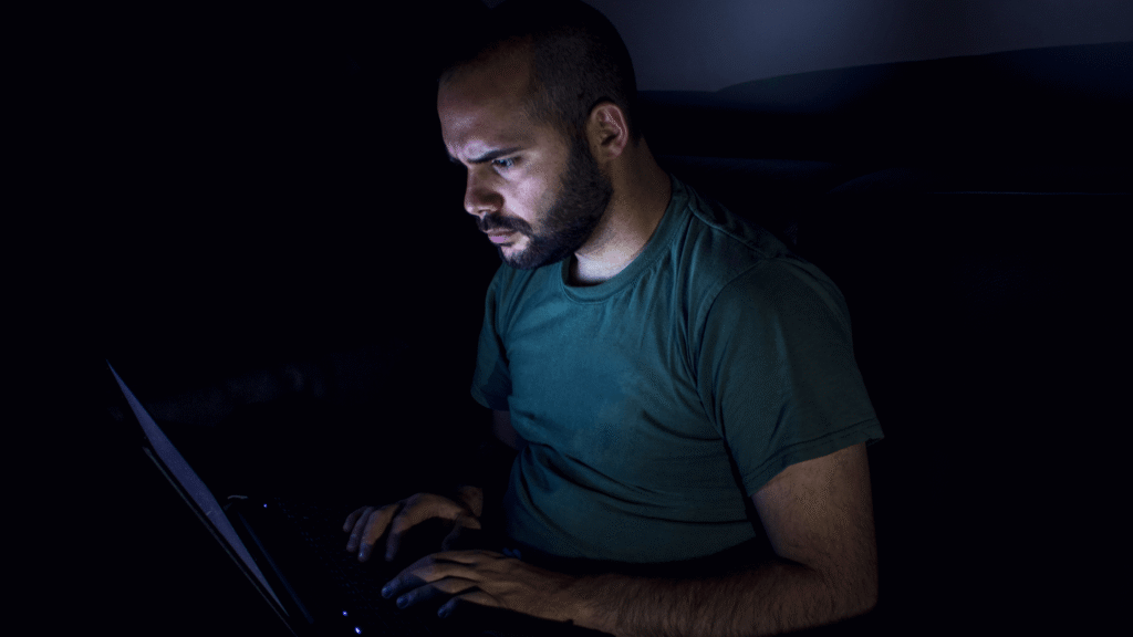 Man sitting on couch in dark room, using laptop on table, face lit by screen, looking serious