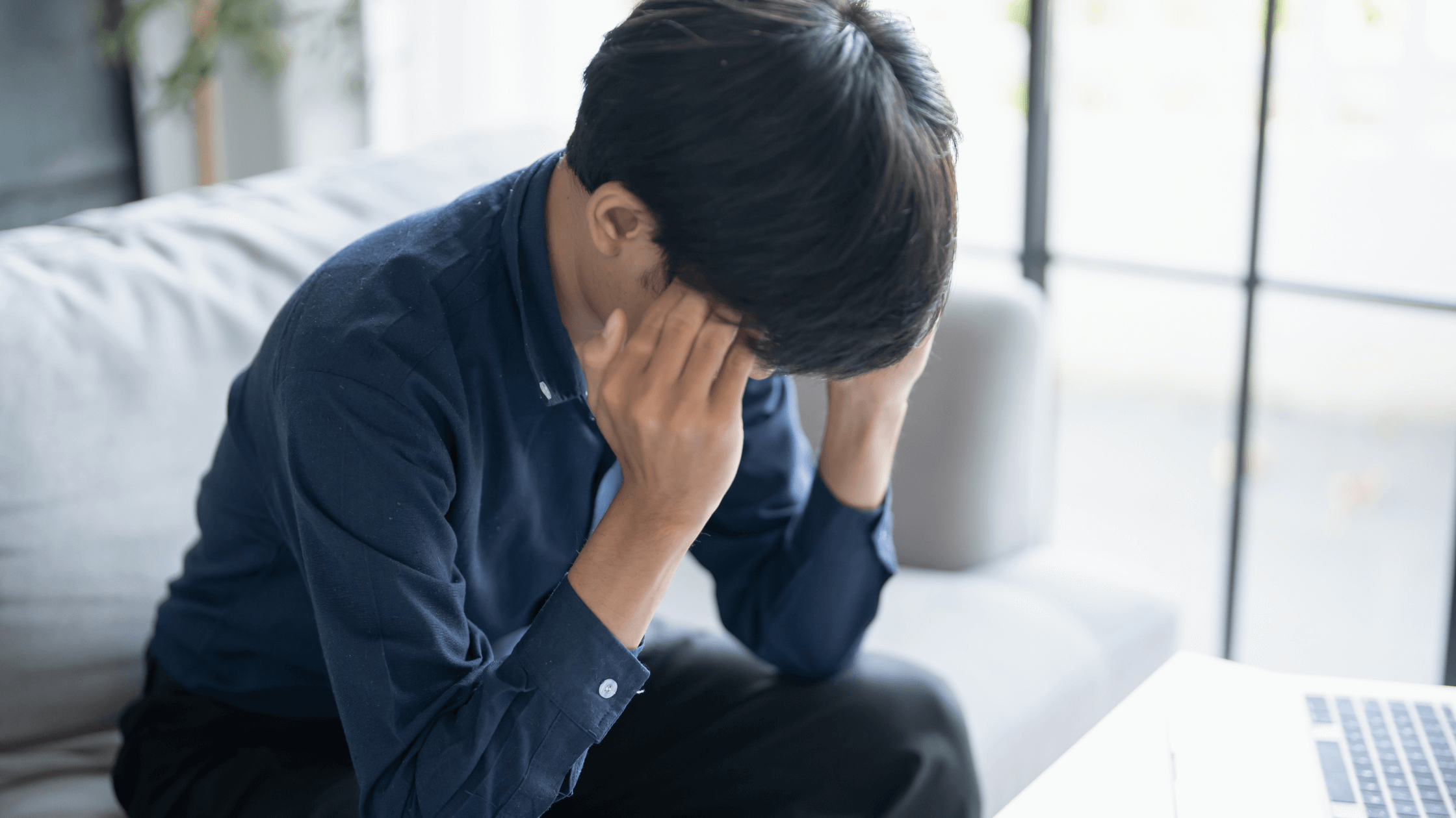 Man seated on couch with laptop on table, hand resting on head, appearing worried