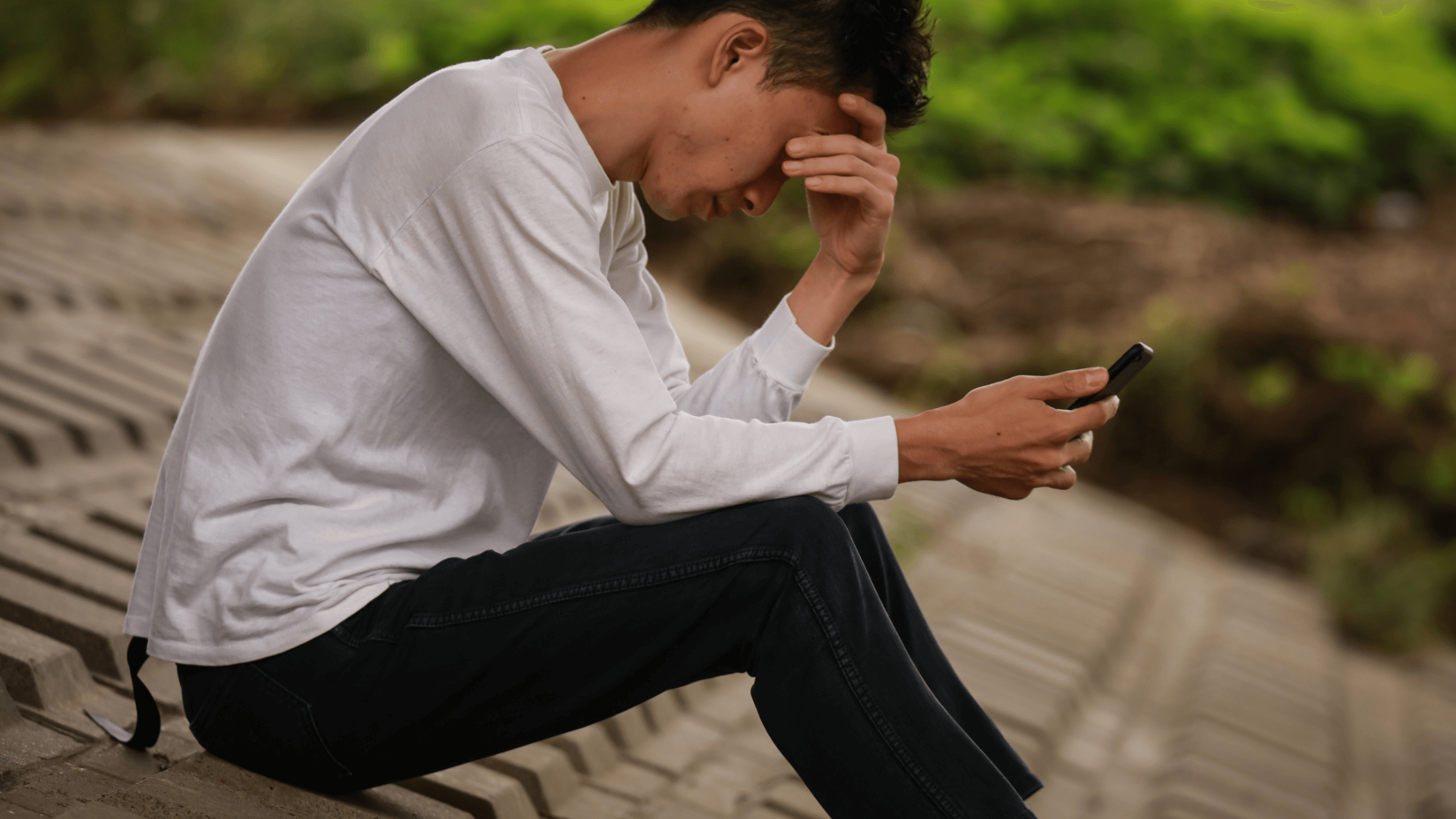 Young man outdoors holding a phone, hand on forehead, appearing sorrowful.