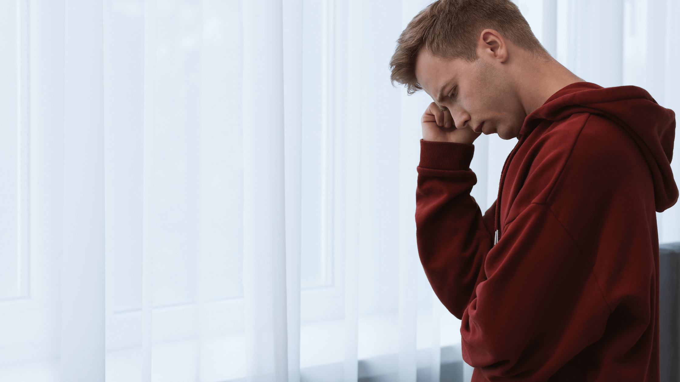 Young man wearing a red sweater standing by the window with a curtain in deep thought.