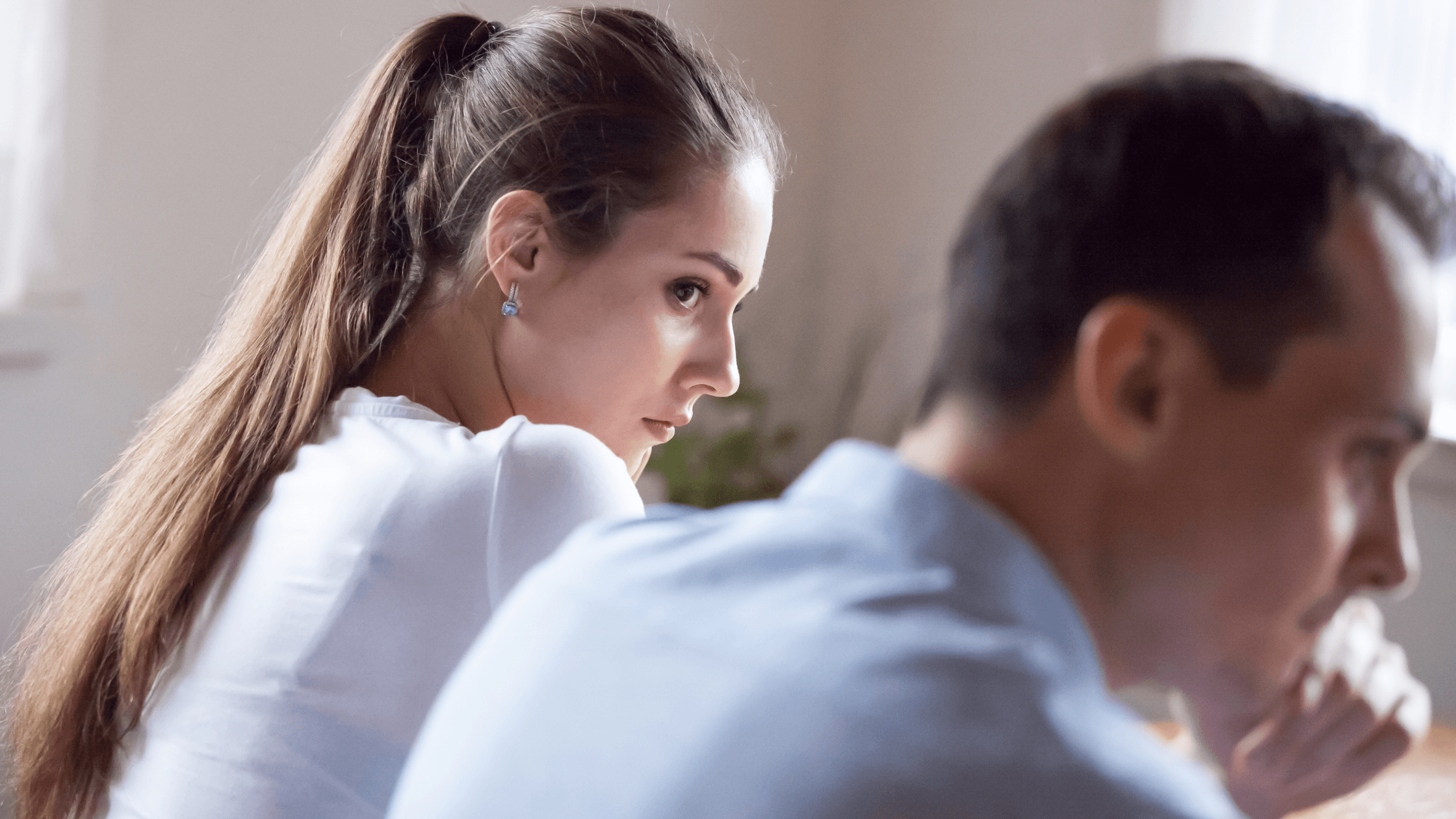 A couple sits on a sofa at home. The woman looks at the man. The man looks away, showing a disagreement.