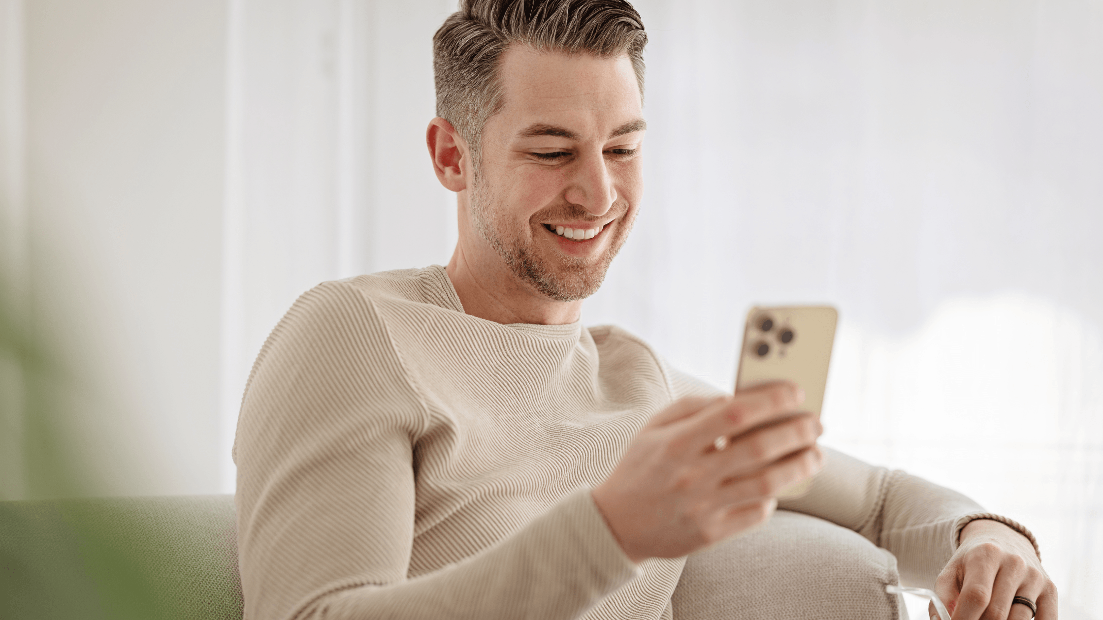 Man sitting comfortably on a couch at home, wearing a sweater, smiling while using a phone.