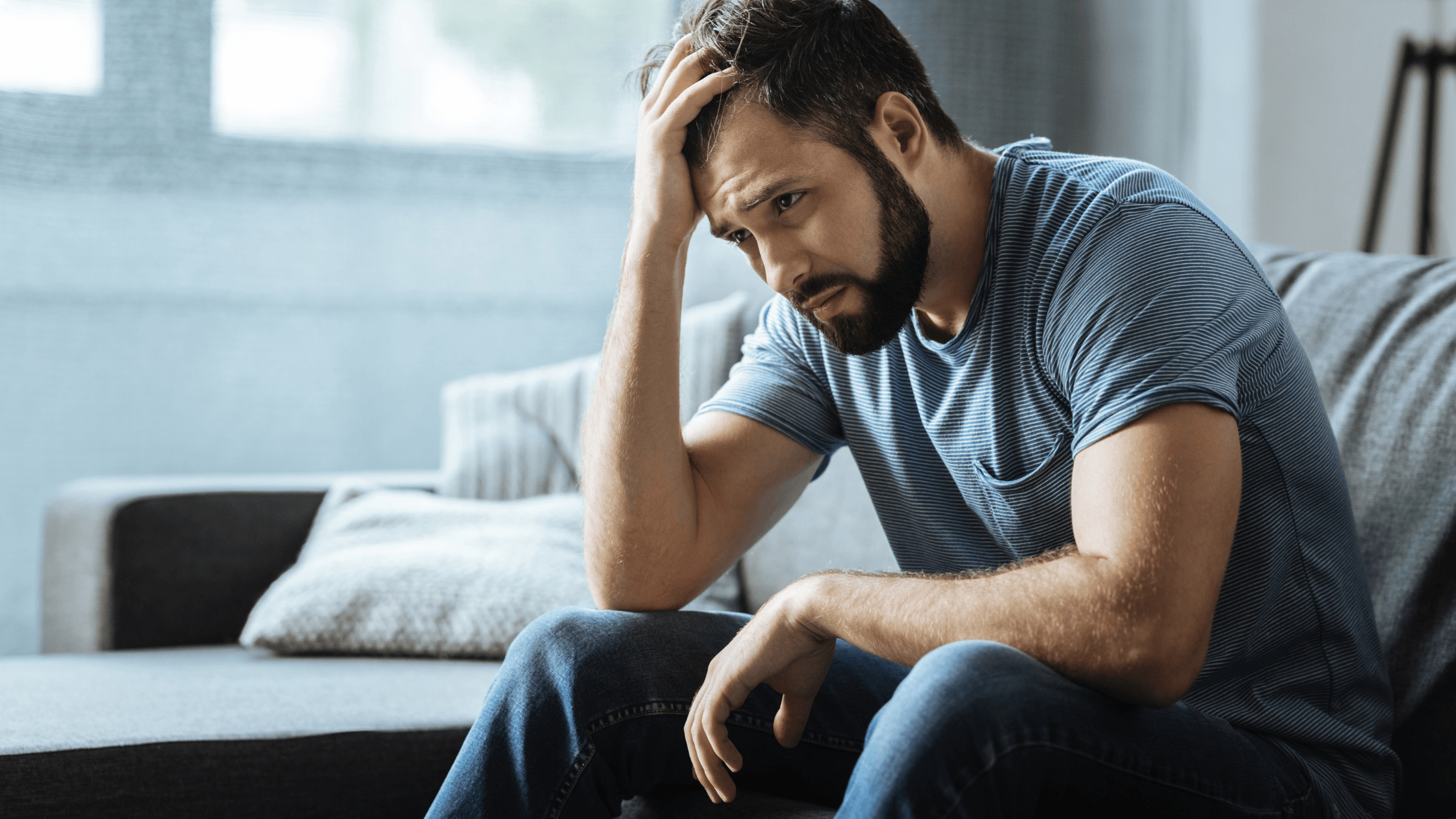 Man sitting in living room with his hand on his head, appearing deep in thought.