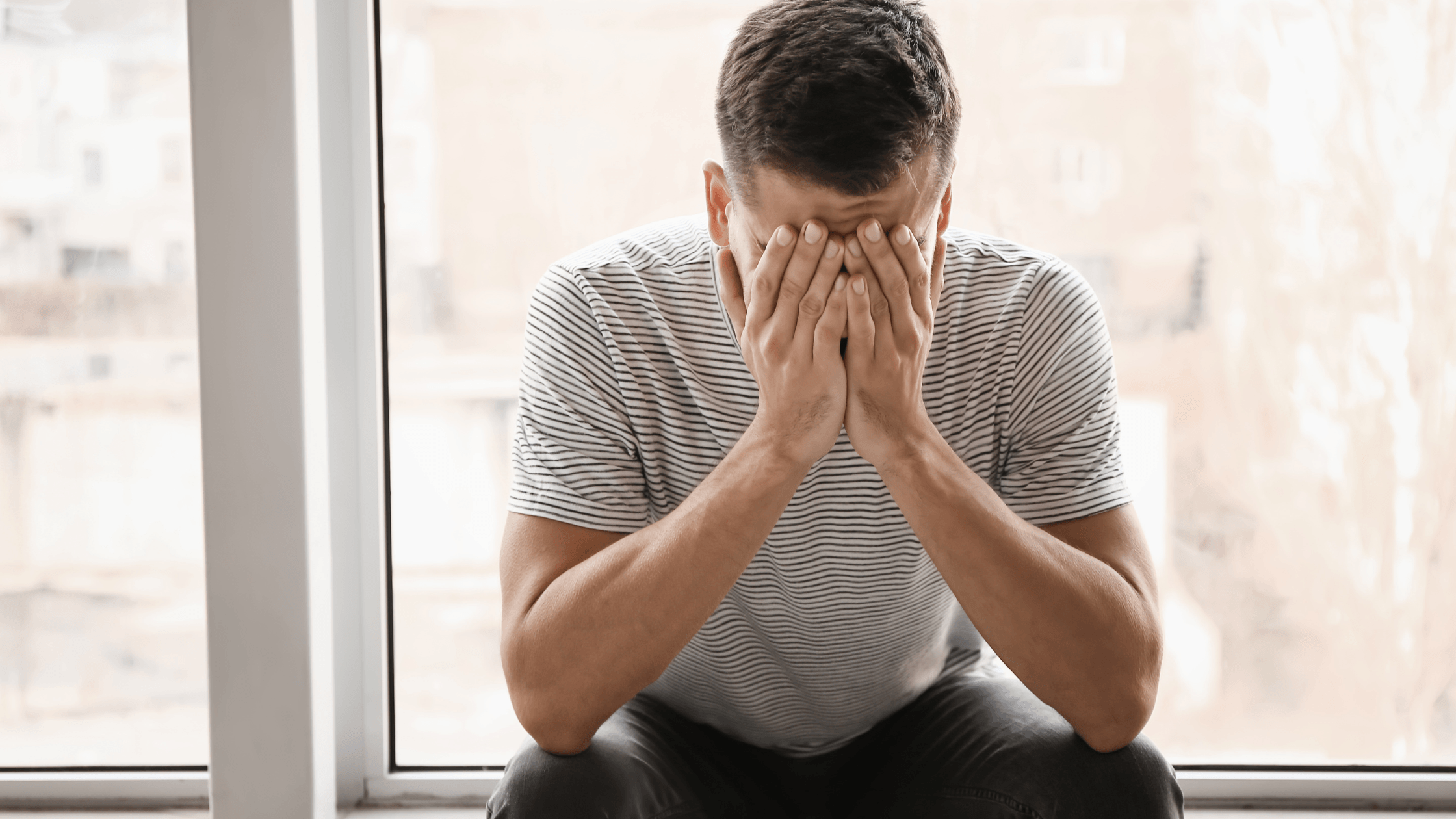 Man alone at home near windows, looking unhappy with his head lowered and hand on his face.