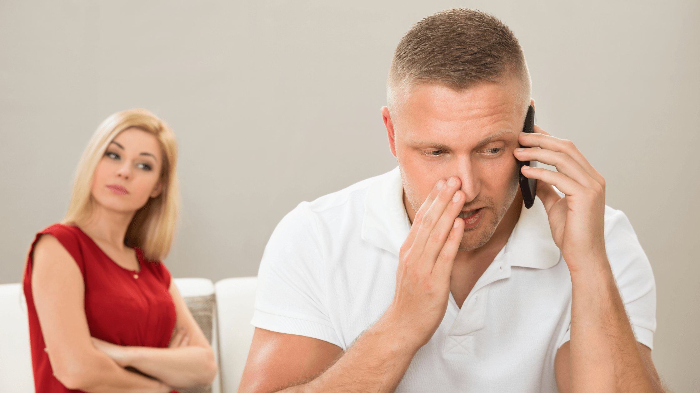 Man on phone call, serious expression, female partner behind him looking suspicious at home
