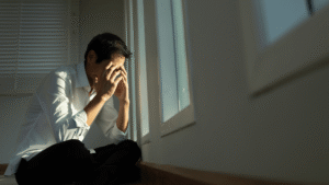 Man sitting on the floor by a window, crying with his hand covering his face.