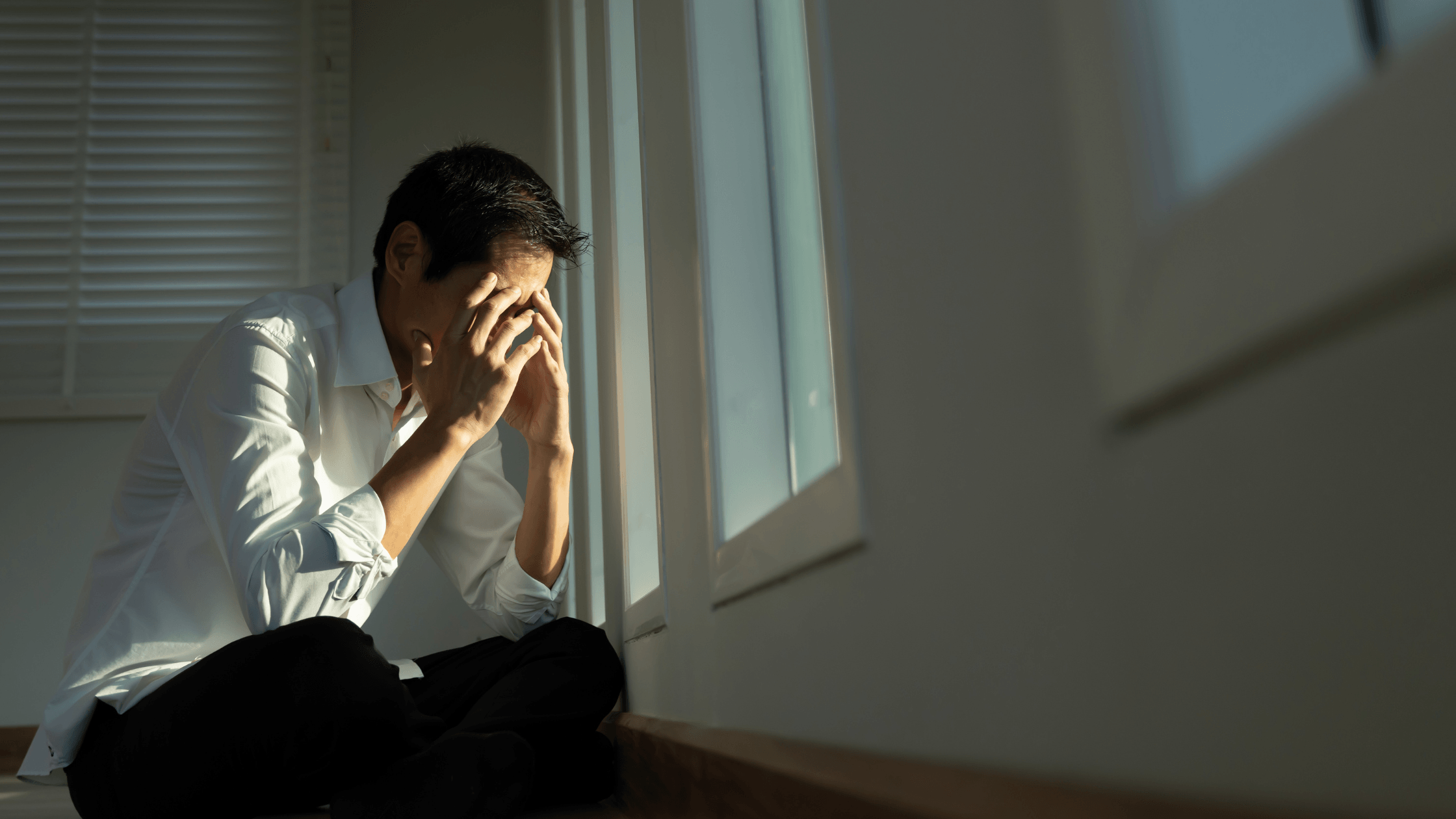 Man sitting on the floor by a window, crying with his hand covering his face.