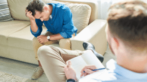 Man sitting on a couch with his hand on his forehead, talking to a therapist who is taking notes.
